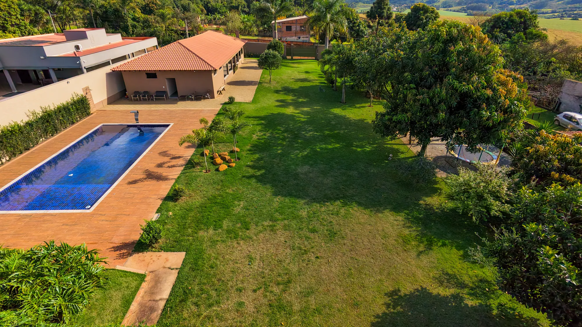 Vista aérea do amplo gramado com árvores, a piscina e o salão ao fundo
