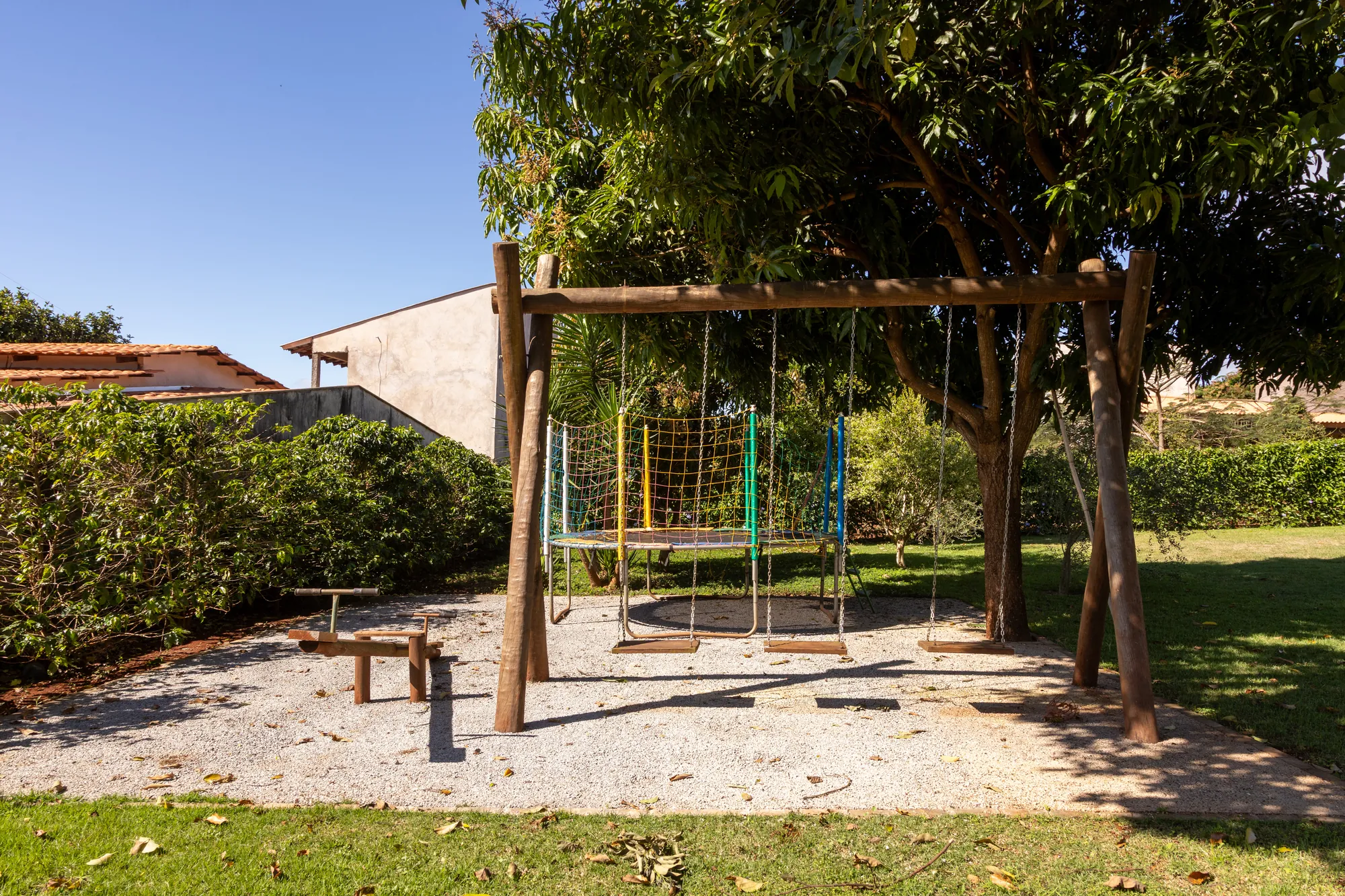 Playground com balanços de madeira e cama elástica sob a sombra de árvores